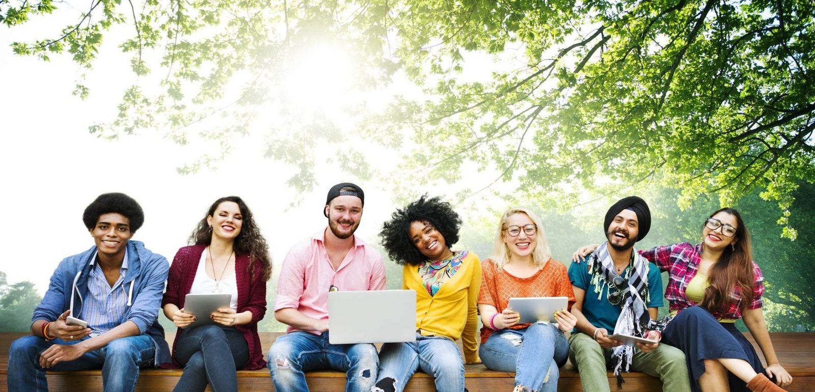 Diverse group of young students smiling and studying outdoors with laptops and tablets under a bright, green canopy of trees.